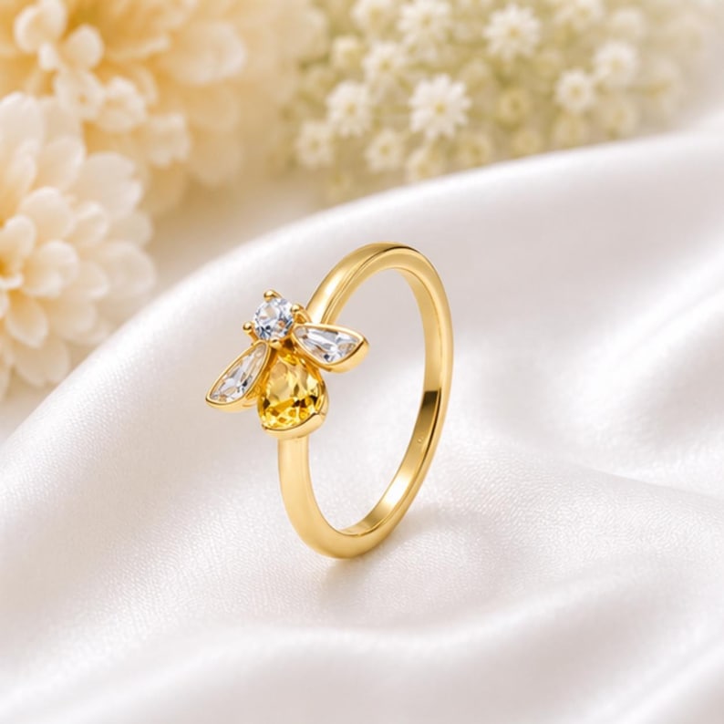 May include: A gold-toned ring featuring a bee-shaped centerpiece. The bee is crafted with a round clear stone for the head, two marquise-cut clear stones for the wings, and a pear-shaped yellow stone for the body, set on a smooth gold band.