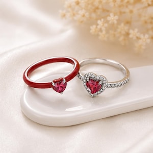 May include: Two rings with heart-shaped gemstones. One ring is red with a matching heart-shaped stone. The other ring is silver with a heart-shaped red stone surrounded by smaller clear stones. Both rings are on a white surface.