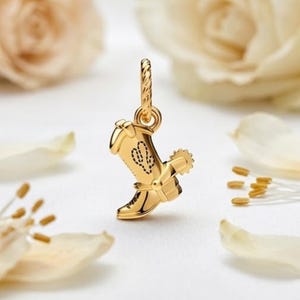 May include: A gold-toned metal charm shaped like a cowboy boot, featuring a small spur on the heel and a dotted cactus design on the side. The charm includes a twisted metal loop at the top for attachment, showcasing a polished, reflective finish.