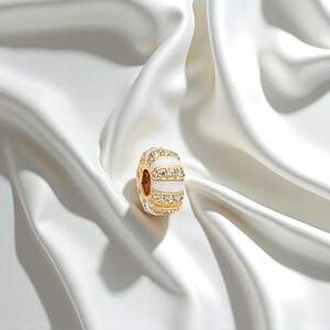 May include: A gold-colored charm with white enamel stripes and rows of small, clear gemstones. The charm is round and has a hole through the center. The background is a white, silky fabric.
