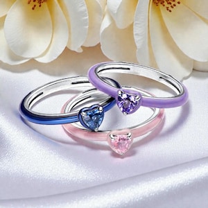 Op de afbeelding: Drie zilveren ringen met hartvormige edelstenen. De ringen hebben blauwe, roze en paarse emaille banden. De edelstenen zijn blauw, roze en paars. De ringen worden gepresenteerd op een witte satijnen ondergrond.