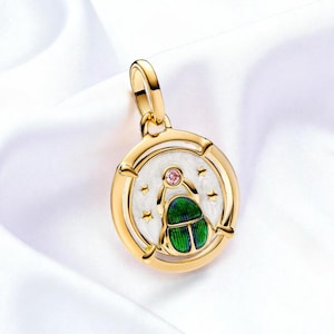 May include: Gold-tone pendant with a circular design featuring a white enamel background, a green and blue scarab beetle, and a pink gemstone. The pendant has a loop for a chain and is set against a white background.