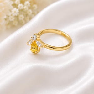 May include: A gold-toned ring featuring a bee-shaped design. The bee has a pear-shaped yellow gemstone for the body, two clear marquise-cut stones for the wings, and a small round clear stone for the head, all set on a smooth, polished gold band.