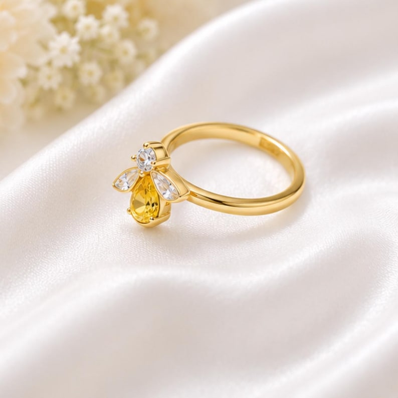 May include: A gold-toned ring featuring a bee-shaped design. The bee has a pear-shaped yellow gemstone for the body, two clear marquise-cut stones for the wings, and a small round clear stone for the head, all set on a smooth, polished gold band.