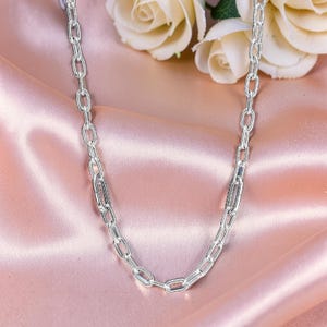 Puede incluir: Un collar de cadena plateada con un diseño de eslabones. El collar se muestra sobre una superficie de satén rosa, con rosas blancas en el fondo. La cadena está hecha de pequeños eslabones rectangulares.