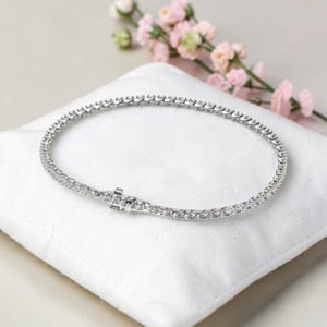 May include: A silver bracelet set with small, clear gemstones. The bracelet is presented on a white cushion, with pink and white flowers in the background. The clasp is clearly visible. The bracelet is likely made of silver.