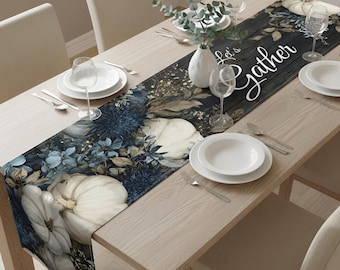 May include: A long table runner featuring a dark wood-grain background with white pumpkins, deep blue floral accents, and the white cursive text Let's Gather. The runner is displayed on a light wood table with white plates, clear glasses, and a vase.