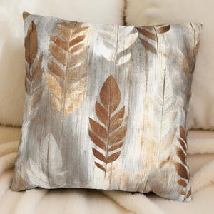 May include: Square throw pillow featuring a repeating pattern of stylized brown and gold feathers on a light gray, wood-grain background. The pillow is square and appears to be made of a soft fabric, suitable for home decor.