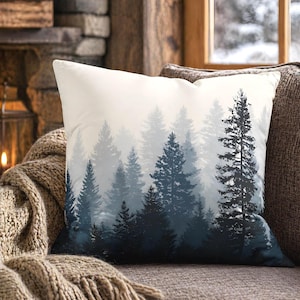 Puede incluir: Un cojín decorativo cuadrado con un estampado de un bosque. El diseño presenta un degradado de árboles de hoja perenne azules y grises, creando un efecto brumoso. El cojín está sobre un sofá marrón con una manta de punto.