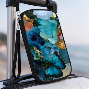 May include: A rectangular black clutch with a vibrant floral and butterfly design. The artwork features shades of blue, gold, and white, with a prominent blue butterfly. Includes a black wrist strap and silver zipper.