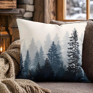 May include: A square throw pillow featuring a printed design of a dense forest with tall pine trees. The color palette transitions from dark navy blue at the bottom to a light, misty white at the top, creating a layered, atmospheric effect.