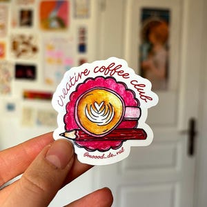 Peut inclure: Un autocollant avec le texte "creative coffee club" en écriture rouge. L'autocollant représente une tasse de café avec du latte art, un crayon rouge et une bordure florale rose. L'autocollant a le compte Instagram @moood_de_val.