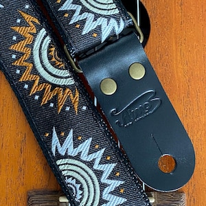 May include: Black guitar strap with a white and gold geometric pattern. The strap has a black leather end with two brass buttons and the word "Vitzo" printed on it.