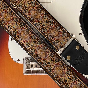 May include: A black guitar strap with a gold and red embroidered pattern. The strap has a leather end with the word "VOLUME" printed on it.