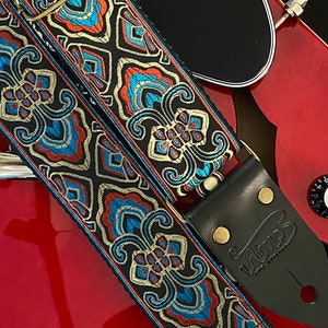 Pode incluir: Uma correia de guitarra preta com um padrão paisley colorido em tons de azul, laranja e dourado. A correia é feita de couro e tem uma fivela de metal. A correia está em uma guitarra vermelha.