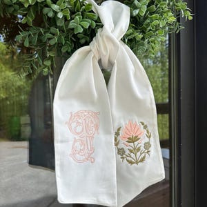 以下が含まれることがあります： 刺繍が施された白いスカーフ。片面にはピンクのモノグラム、もう片面にはグリーンのアクセントが付いたピンクの花柄のデザインがあります。スカーフは結ばれており、緑のリースに掛けられています。