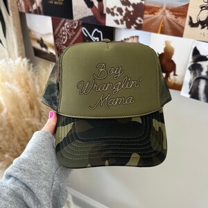 May include: A green camouflage trucker hat with a brown embroidered design that says "Boy Wranglin' Mama".