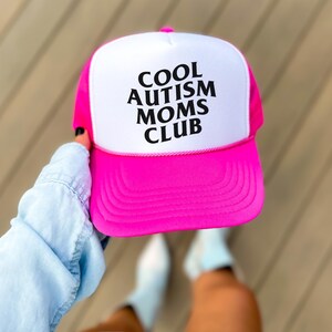 May include: A pink and white trucker hat with the text "COOL AUTISM MOMS CLUB" in black lettering.