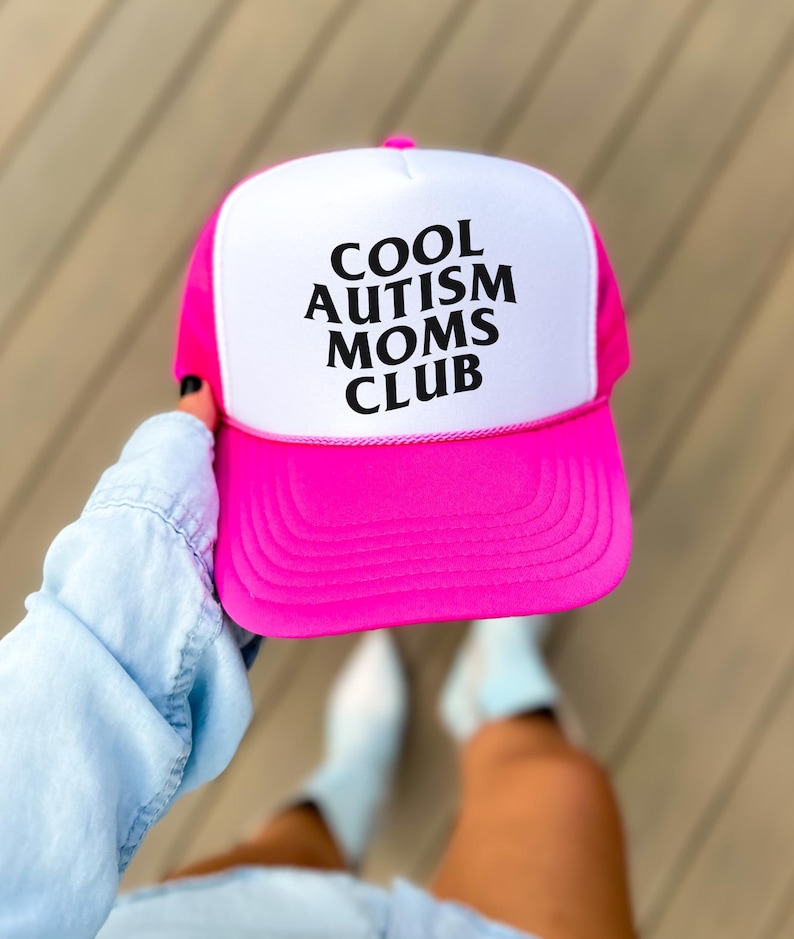 May include: A pink and white trucker hat with the text "COOL AUTISM MOMS CLUB" in black lettering.