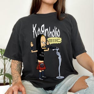 May include: A black t-shirt with a cartoon image of a person wearing a kilt and holding a microphone. The text "Kornholio" is printed above the image and the text "BOOM NA DA BOOM NOOM NA NEMA BOOM NA DA BOOM!" is printed below the image. The text "NU METAL" is printed on the person's shirt.