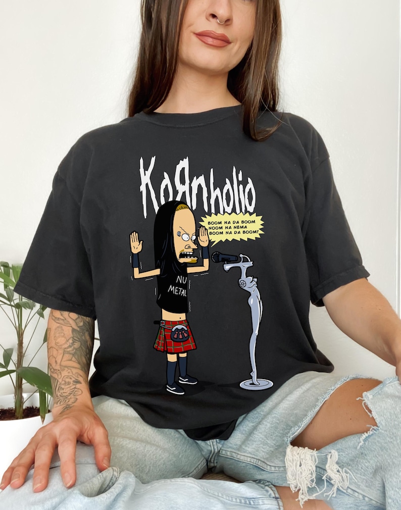 May include: A black t-shirt with a cartoon image of a person wearing a kilt and holding a microphone. The text "Kornholio" is printed above the image and the text "BOOM NA DA BOOM NOOM NA NEMA BOOM NA DA BOOM!" is printed below the image. The text "NU METAL" is printed on the person's shirt.