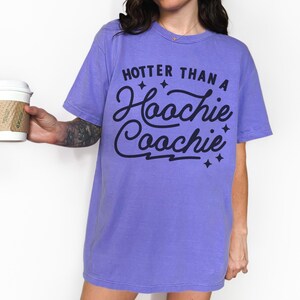 Puede incluir: Una camiseta de color lila con la frase "Hotter Than a Hoochie Coochie" en negro. La camiseta tiene un corte relajado y mangas cortas. El texto est&aacute; en una fuente cursiva juguetona con estrellas.