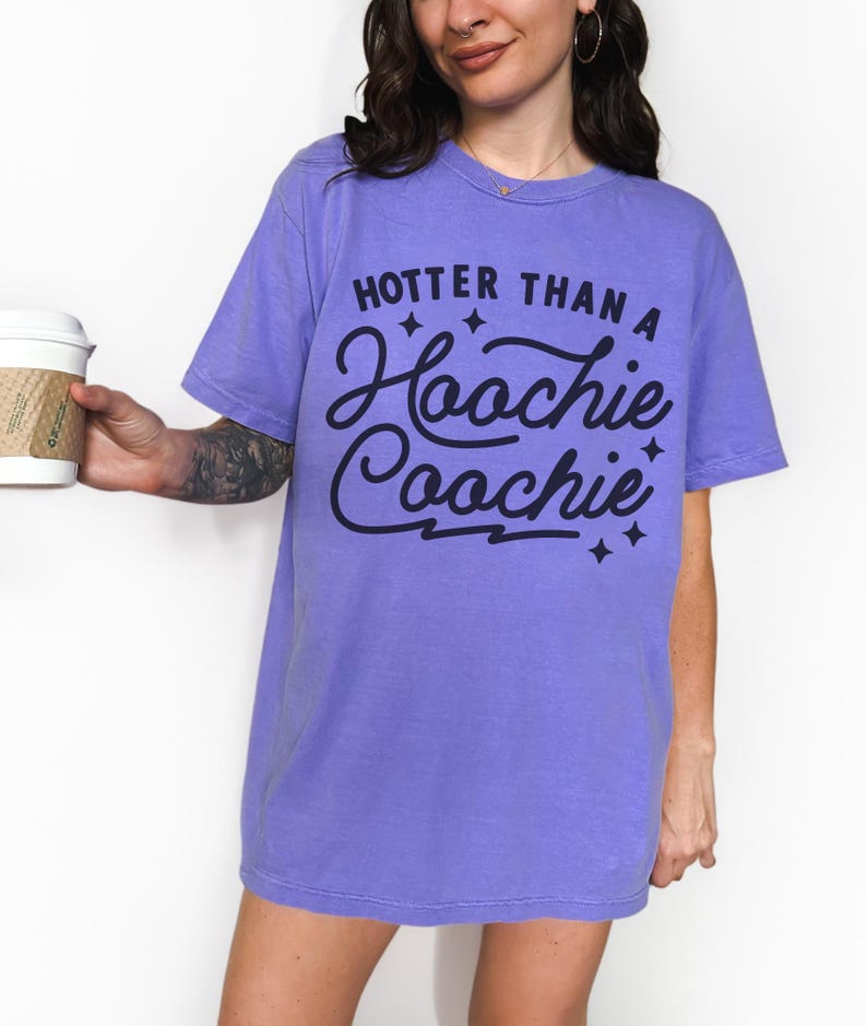 Puede incluir: Una camiseta de color lila con la frase "Hotter Than a Hoochie Coochie" en negro. La camiseta tiene un corte relajado y mangas cortas. El texto est&aacute; en una fuente cursiva juguetona con estrellas.
