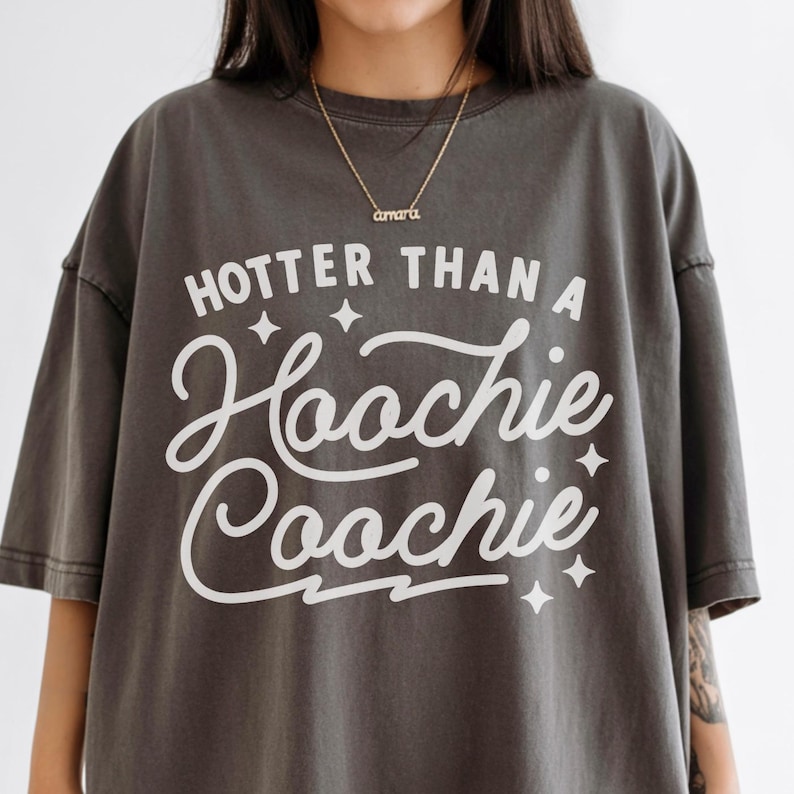 Puede incluir: Camiseta gris oscuro con la frase blanca "HOTTER THAN A Hoochie Coochie". La camiseta tiene un corte holgado y mangas cortas. Se ve un collar dorado con el nombre "amara".