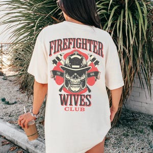 May include: Cream-colored t-shirt with a graphic on the back featuring a firefighter's helmet, skull, and crossed axes. The text "FIREFIGHTER WIVES CLUB" is printed in red and black.