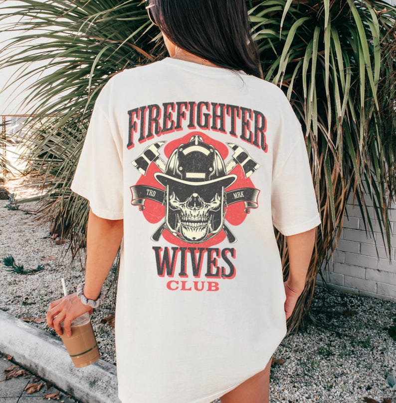 May include: Cream-colored t-shirt with a graphic on the back featuring a firefighter's helmet, skull, and crossed axes. The text "FIREFIGHTER WIVES CLUB" is printed in red and black.