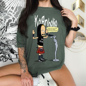 May include: A green t-shirt with a cartoon image of a person with black hair and a red kilt. The person is holding a microphone and the text "Kornholio" is printed above the image. The text "BOOM NA DA BOOM NOOM NA NEMA BOOM NA DA BOOM!" is printed below the image. The text "NU METAL" is printed on the person's shirt.