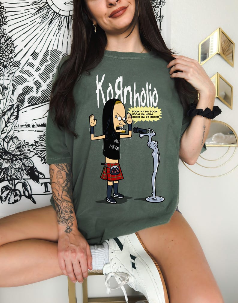 May include: A green t-shirt with a cartoon image of a person with black hair and a red kilt. The person is holding a microphone and the text "Kornholio" is printed above the image. The text "BOOM NA DA BOOM NOOM NA NEMA BOOM NA DA BOOM!" is printed below the image. The text "NU METAL" is printed on the person's shirt.