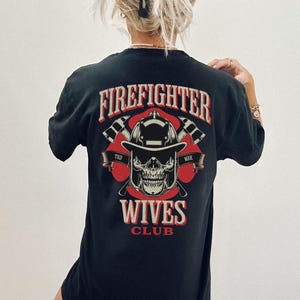 May include: Black t-shirt with a graphic design featuring the text "FIREFIGHTER" above a skull wearing a firefighter helmet, crossed axes, and the words "WIVES CLUB". The design is in red and white.
