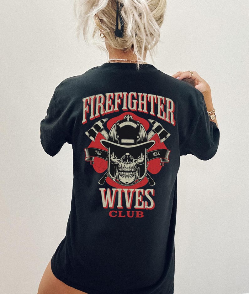 May include: Black t-shirt with a graphic design featuring the text "FIREFIGHTER" above a skull wearing a firefighter helmet, crossed axes, and the words "WIVES CLUB". The design is in red and white.