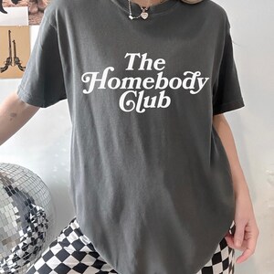Może przedstawiać: Szary T-shirt z napisem "The Homebody Club" w białym, ozdobnym pismem.