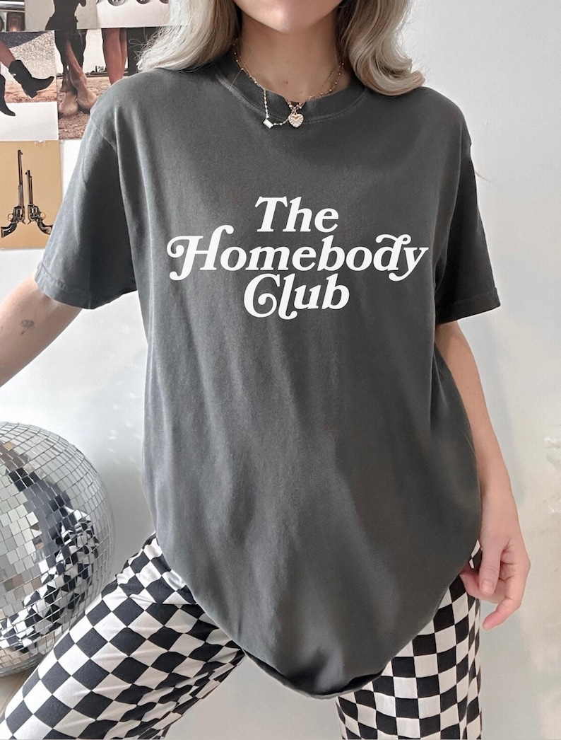 Może przedstawiać: Szary T-shirt z napisem "The Homebody Club" w białym, ozdobnym pismem.