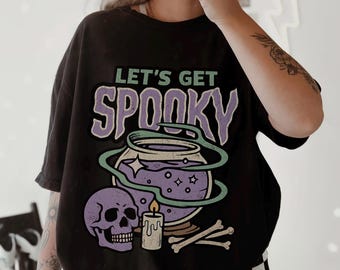 May include: A black short-sleeve t-shirt featuring a graphic print with the text "LET'S GET SPOOKY" in green and purple. The design includes a purple skull, a lit candle, a cauldron with purple liquid, and bones, all outlined in green.
