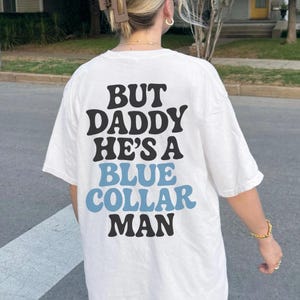 May include: White t-shirt with the text "BUT DADDY HE'S A BLUE COLLAR MAN" in black and light blue lettering. The shirt is a casual style.