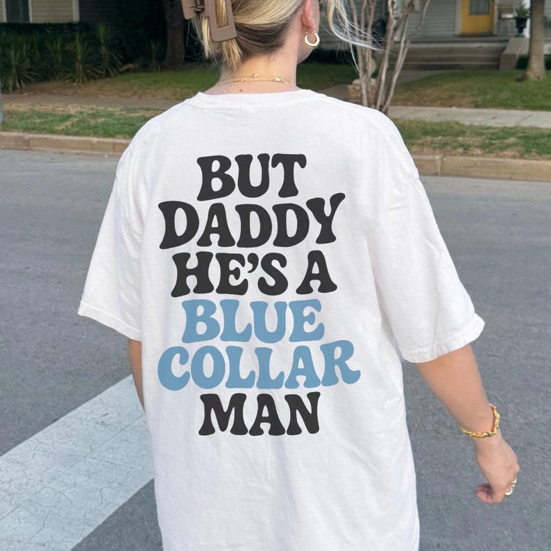 May include: White t-shirt with the text "BUT DADDY HE'S A BLUE COLLAR MAN" in black and light blue lettering. The shirt is a casual style.