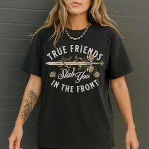 Pode incluir: T-shirt cinzento escuro com o texto "TRUE FRIENDS Stab You IN THE FRONT" em estilo vintage. O design inclui uma espada, rosas e elementos decorativos. Uma peça de roupa casual.