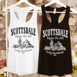 May include: Two tank tops, one white and one black, hanging on a white hanger. Each tank top has the text "SCOTTSDALE Before The Veil" with a desert landscape graphic and "Emily's Bachelorette 2025".