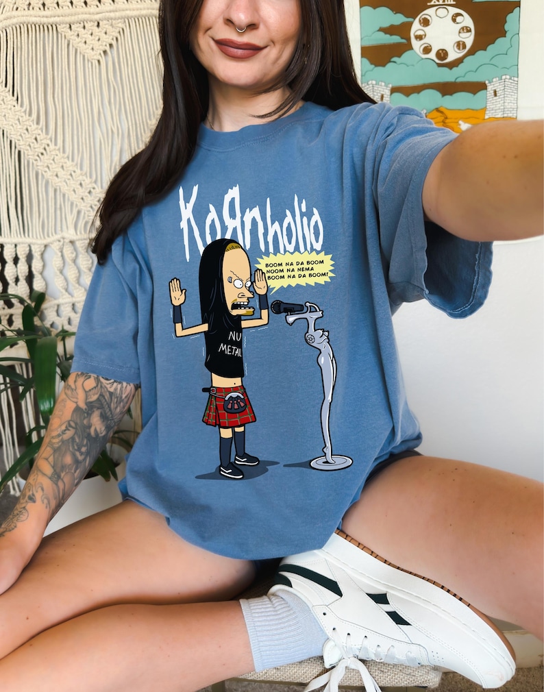 May include: A blue t-shirt with a cartoon image of a character wearing a kilt and holding a microphone. The text "Kaanholio" is printed on the shirt, along with the phrase "BOOM NA DA BOOM NOOM HA NEMA BOOM NA DA BOOM!" and "NU METAL".