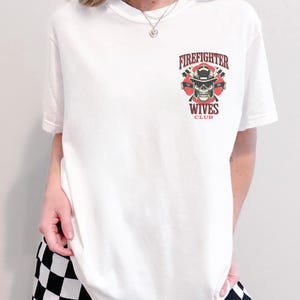 May include: White t-shirt featuring a graphic with the text "Firefighter Wives Club". The graphic includes a skull wearing a firefighter helmet, crossed axes, and the words "Firefighter" and "Wives Club" in red and black.