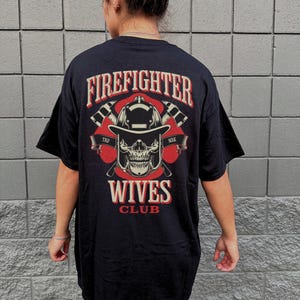 May include: Black t-shirt with a graphic on the back featuring a firefighter skull wearing a helmet, crossed axes, and the words "Firefighter Wives Club" in red and white. The shirt is worn by a person.