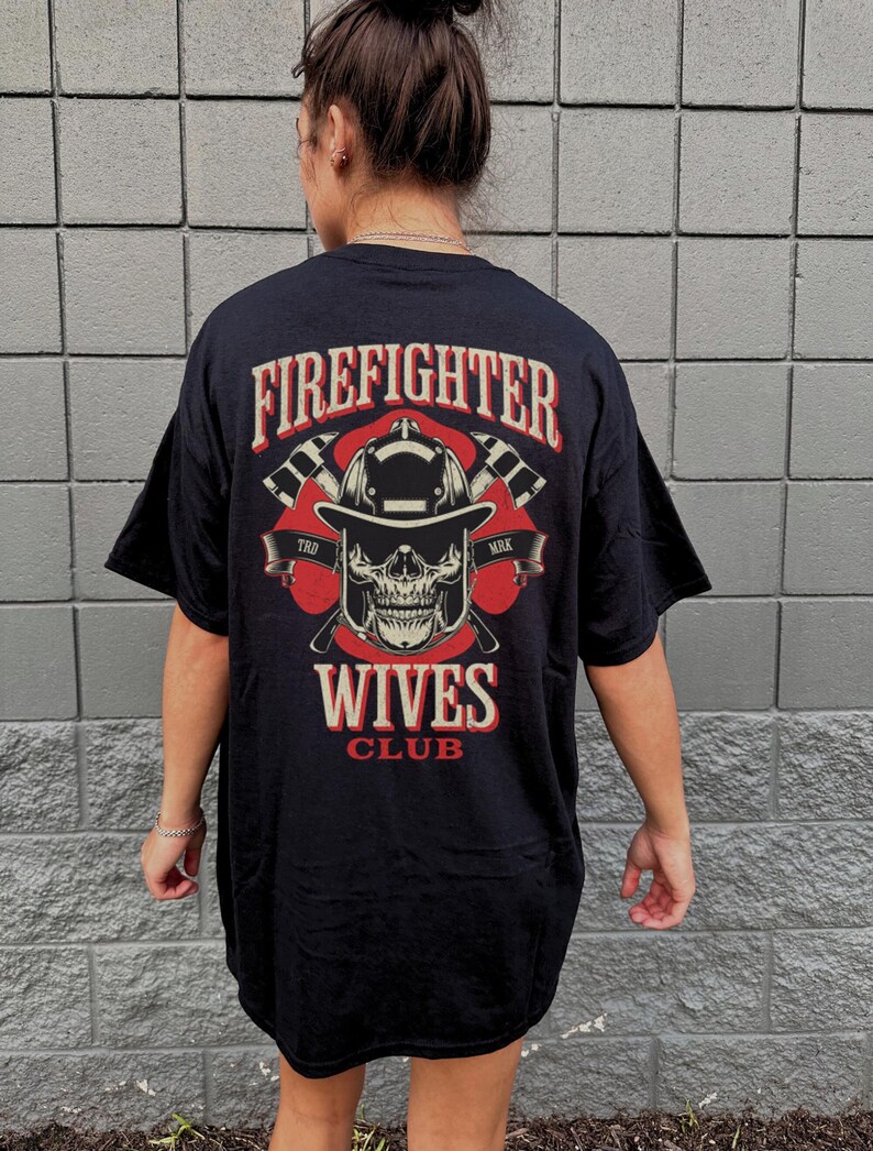 May include: Black t-shirt with a graphic on the back featuring a firefighter skull wearing a helmet, crossed axes, and the words "Firefighter Wives Club" in red and white. The shirt is worn by a person.