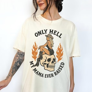 May include: Cream-colored t-shirt featuring a graphic of a woman in a cowboy hat sitting on a skull, surrounded by flames. The text reads "ONLY HELL MY MAMA EVER RAISED" in a circular design. The shirt has short sleeves.