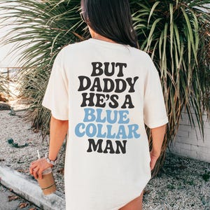 May include: Cream-colored t-shirt with the text "BUT DADDY HE'S A BLUE COLLAR MAN" in black and blue lettering. The shirt is worn by a person standing in front of green foliage.