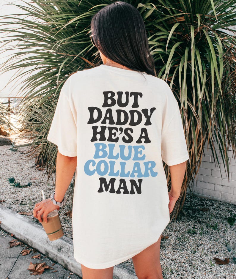 May include: Cream-colored t-shirt with the text "BUT DADDY HE'S A BLUE COLLAR MAN" in black and blue lettering. The shirt is worn by a person standing in front of green foliage.