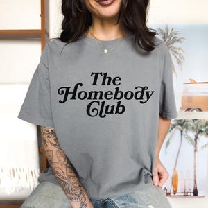 Może przedstawiać: Szary T-shirt z napisem "The Homebody Club" czarnymi literami.