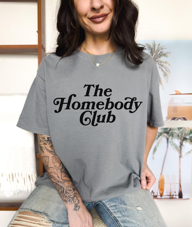 Może przedstawiać: Szary T-shirt z napisem "The Homebody Club" czarnymi literami.